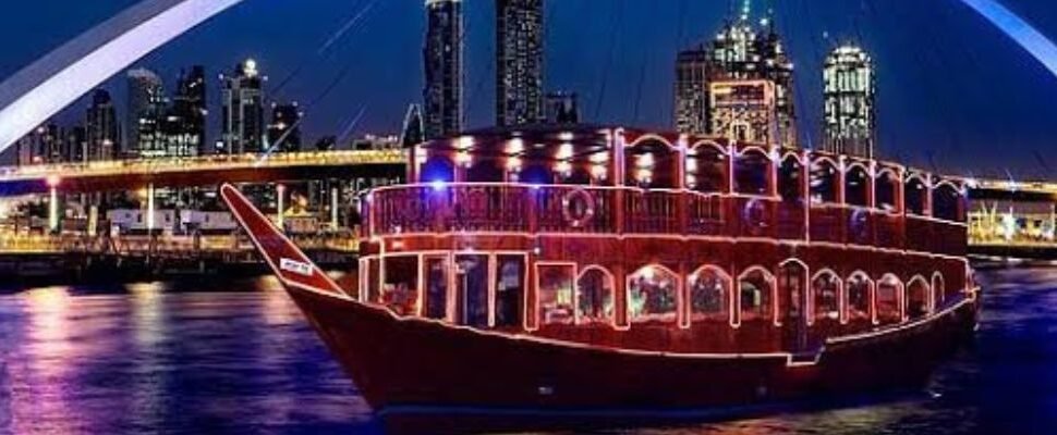 A traditional dhow boat sails on the serene waters of Dubai at night, with the city skyline illuminated in the background.