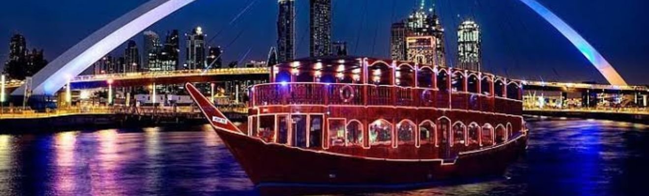 A traditional dhow boat sails on the serene waters of Dubai at night, with the city skyline illuminated in the background.