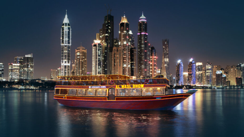A brightly illuminated dhow cruise glides along calm waters at night, with the glowing skyscrapers of a modern city skyline towering in the background, highlighting the charm of the Creek Dhow Cruise Dubai, the cultural appeal seen in Dhow Cruise Dubai Creek vs Marina, the scenic beauty of the Dhow Cruise Dubai Creek location, the popularity of the Best Dhow Cruise Dubai Creek, the convenience of flexible Dhow Cruise Dubai Creek timings and Dhow Cruise Creek timings, and offering a striking contrast to the vibrant Dhow Cruise Dubai Marina experience.