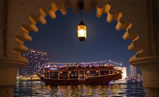 A dhow cruise boat on a night-time waterfront framed artistically by an ornate archway with a hanging lantern, capturing the charm of the Dubai Creek Dhow Cruise, showcasing the appeal of the Best Dhow Cruise Dubai Creek, and reflecting the convenience of flexible Dhow Cruise Dubai Creek timings and Dhow Cruise Creek timings, while offering a beautiful contrast to the modern experience of the Dhow Cruise Dubai Marina.