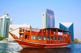 A traditional wooden dhow boat sails on a calm waterway with modern high-rise buildings in the background on a bright, clear day, showcasing the beauty of the Creek Dhow Cruise Dubai, while highlighting the difference seen in Dhow Cruise Dubai Creek vs Marina, the scenic charm of the Dhow Cruise Dubai Creek location, the appeal of the Best Dhow Cruise Dubai Creek, and the convenience offered by flexible Dhow Cruise Dubai Creek timings.