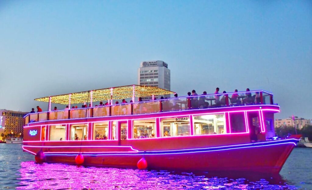 A brightly lit wooden dhow cruise glides along the water at night, with passengers dining inside and Dubai’s illuminated skyscrapers rising in the background, capturing the charm of the Dubai Creek Dhow Cruise and the scenic Dhow Cruise Dubai Creek location, showcasing the appeal of the Best Dhow Cruise Dubai Creek, the ease of Dhow Cruise Dubai Creek timings and Dhow Cruise Creek timings, while offering a striking contrast to the modern experience of the Dhow Cruise Dubai Marina.