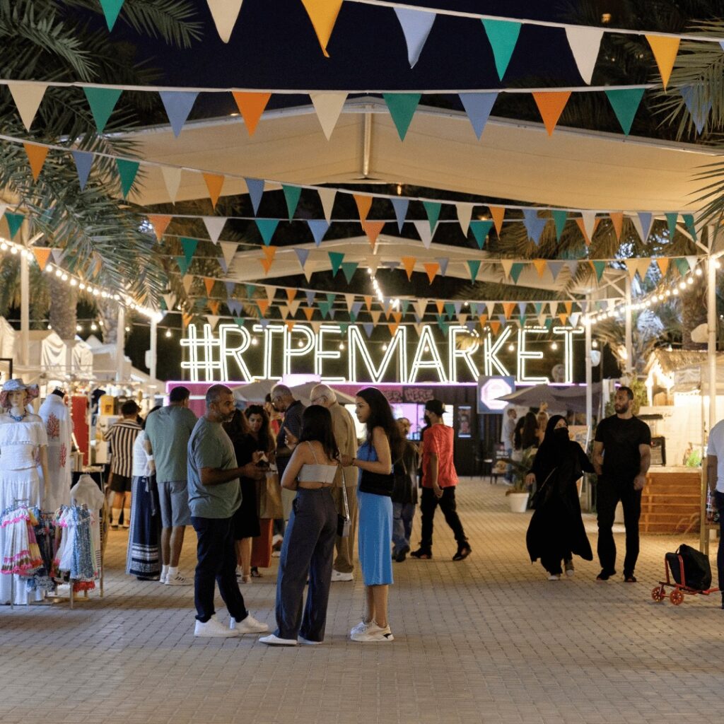 A bustling Dubai night markets under colorful flags and fairy lights, with people shopping, chatting, and enjoying the vibrant atmosphere at #RipeMarket.