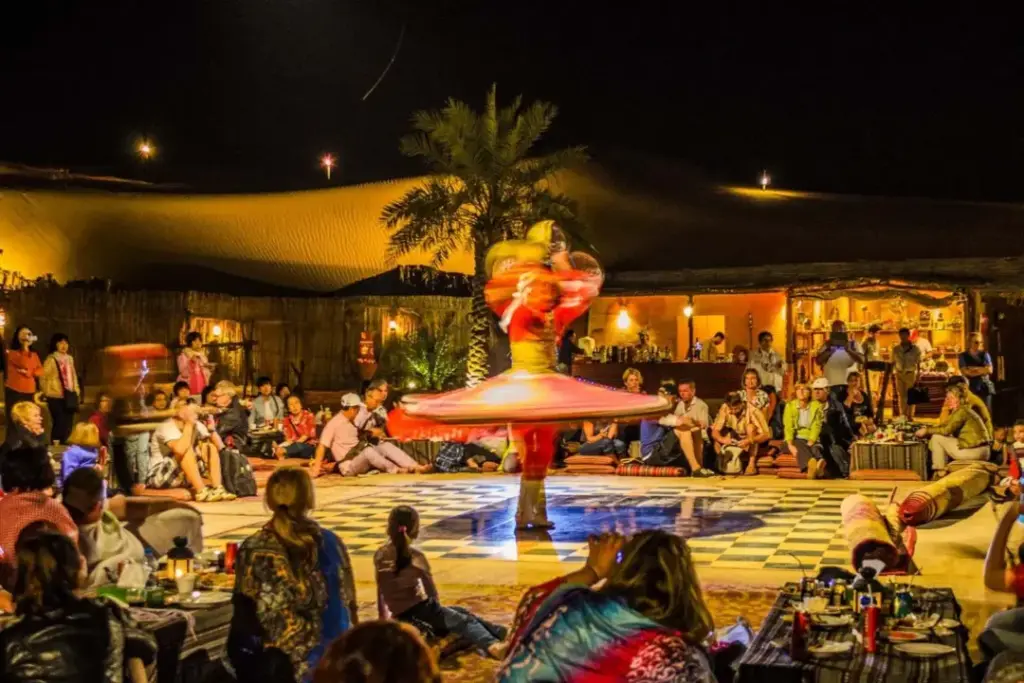 A lively evening in Dubai with tourists enjoying a tanoura dance performance under the stars, surrounded by lanterns and sand dunes near traditional night markets.
