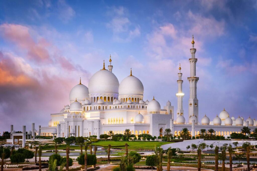 A majestic white mosque with large domes and tall minarets illuminated at dusk, surrounded by lush gardens and palm trees under a colorful sky. This beautiful scene reflects the elegance and grandeur often featured in a Dubai city half day sightseeing tour. The image embodies the essence of a half day Dubai city tour inclusions, offering a glimpse of Dubai’s rich architectural beauty. For a more comprehensive experience, consider the Dubai half day city tour package, which includes visits to iconic landmarks. The Dubai half day city tour timings are typically set to allow for a relaxed and informative journey. For added excitement, opt for a Dubai half day city tour with monorail ride to see the city from a different perspective. This tour's half day Dubai city tour includes convenient transport and expert guides to help you explore the best of Dubai in a short time.
