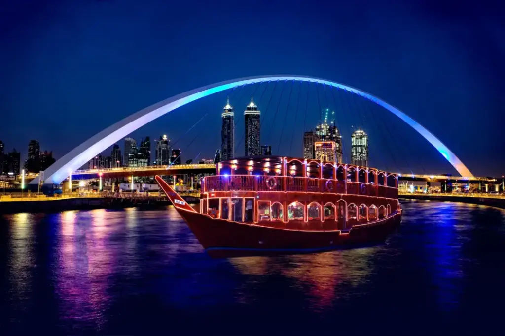 Canal_Dinner › Forever Tourism LLC A brightly lit boat sailing at night with a glowing blue arch bridge and a city skyline in the background, capturing the charm of the Dubai Canal Dhow Cruise, the elegance of the Luxury Canal Dinner Cruise Dubai, the peaceful experience of the Canal Bay Dinner Cruise Dubai, the value offered through Dubai Canal Dhow Cruise offers, and the scenic beauty of the Dubai Water Canal Dhow Cruise.