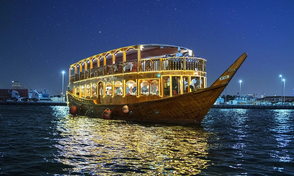 Traditional wooden boat sailing on a Dhow Cruise Dubai in Marina, featuring a Dubai dinner cruise with illuminated city skyline in the background.