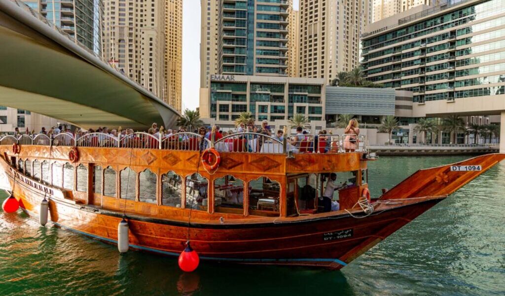 Traditional wooden cruise ship sailing on Dubai Creek during a Dhow Cruise in Dubai with guests enjoying a buffet dinner on deck.