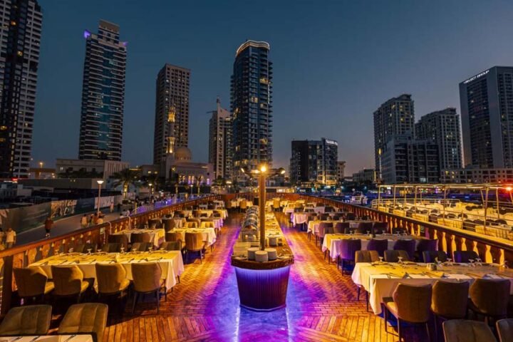 Marina Dhow Cruise: #1 Evening Tour In Dubai! › Forever Tourism LLC Traditional wooden boat sailing on Dubai Marina at sunset during a Marina Dhow Cruise, showcasing skyline views and onboard dining.