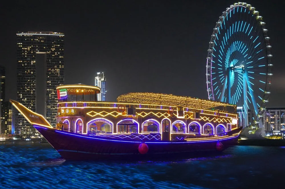 Traditional wooden dhow sailing through Dubai Marina at night, offering a Dhow Cruise in Dubai with dinner, city lights, and scenic waterfront views.