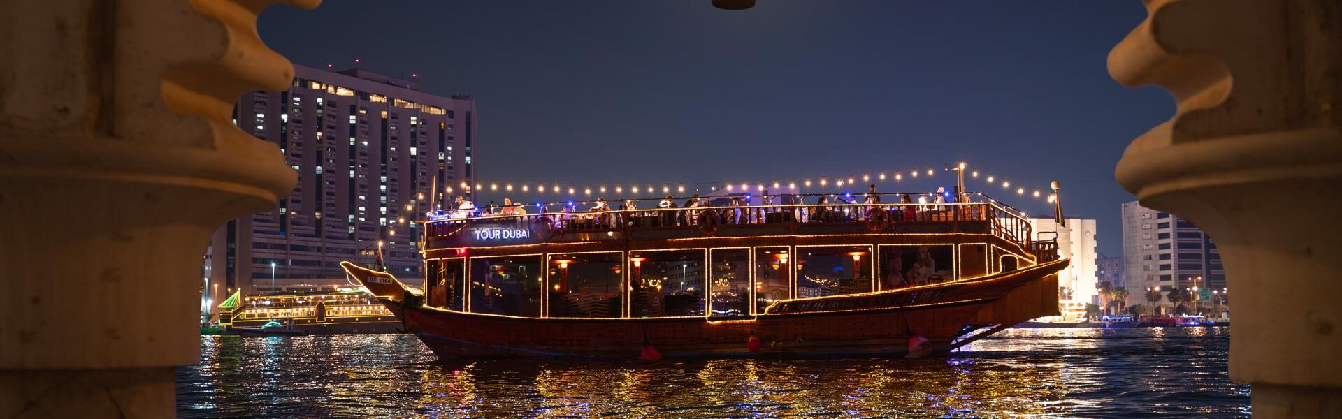 Traditional wooden boat on a Dubai Creek Dhow Cruise with guests enjoying a Dhow Dinner Cruise along the Dhow Cruise Creek at sunset.