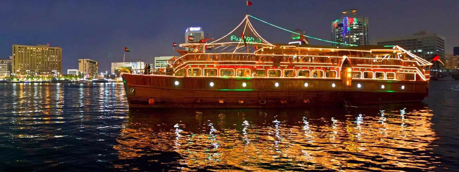 Traditional wooden dhow sailing on Dubai Creek during a Creek Dhow Cruise Dinner with guests enjoying a buffet and night views.