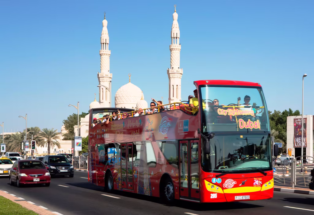 Red Dubai city tour bus with tourists enjoying a hop-on hop-off sightseeing experience in front of iconic Dubai landmarks.