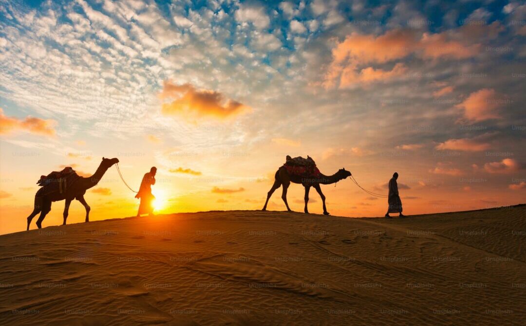 Sunset Camel Caravan — dubai desert photos › Forever Tourism LLC dubai desert photos: silhouettes of two guides leading camels across rippled dunes at sunset beneath a cloud-streaked sky.