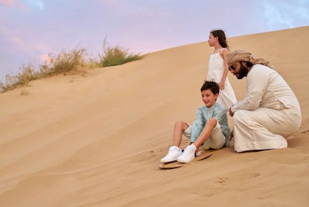 Sandboarding Dubai — boy sliding down a gentle dune on a seated board with a guide and sister at sunset.