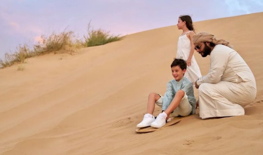 Sandboarding Dubai — boy sliding down a gentle dune on a seated board with a guide and sister at sunset.