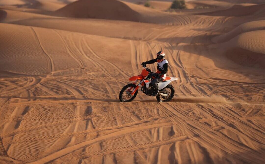 Dirt Bike Dubai – Desert Adventure Ride › Forever Tourism LLC A rider on a red dirt bike speeding across golden desert dunes in Dubai during sunset.