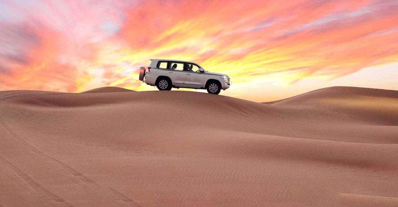 Dubai Desert View — Sunset 4x4 on Golden Dunes › Forever Tourism LLC Dubai Desert View at sunset with a white 4x4 on rolling golden dunes beneath a vibrant pink-orange sky.