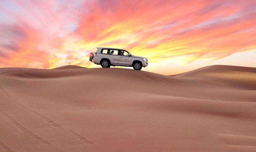 Dubai Desert View at sunset with a white 4x4 on rolling golden dunes beneath a vibrant pink-orange sky.