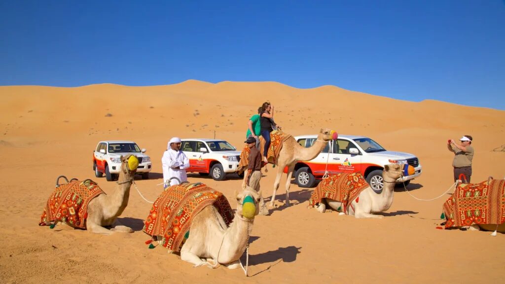 4x4 cresting red dunes at sunset during an Adventure Safari Dubai, with a sandboarder in the foreground and a Bedouin-style camp in the distance.