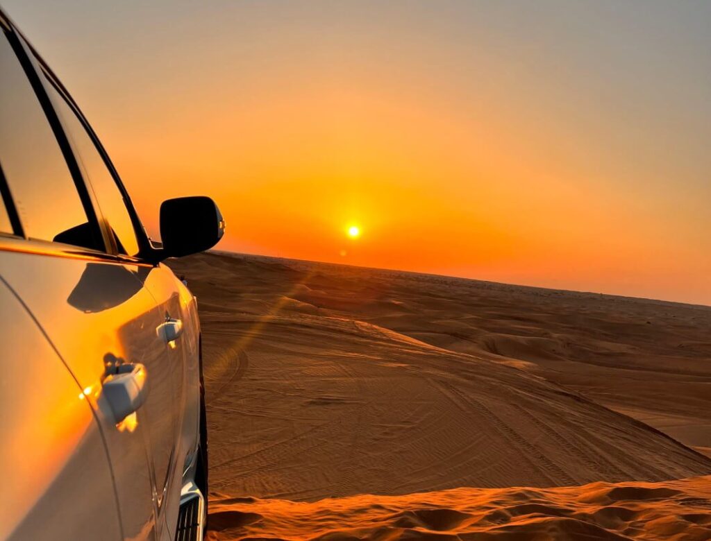 4x4 cresting red dunes at sunset during an Adventure Safari Dubai, with a sandboarder in the foreground and a Bedouin-style camp in the distance.