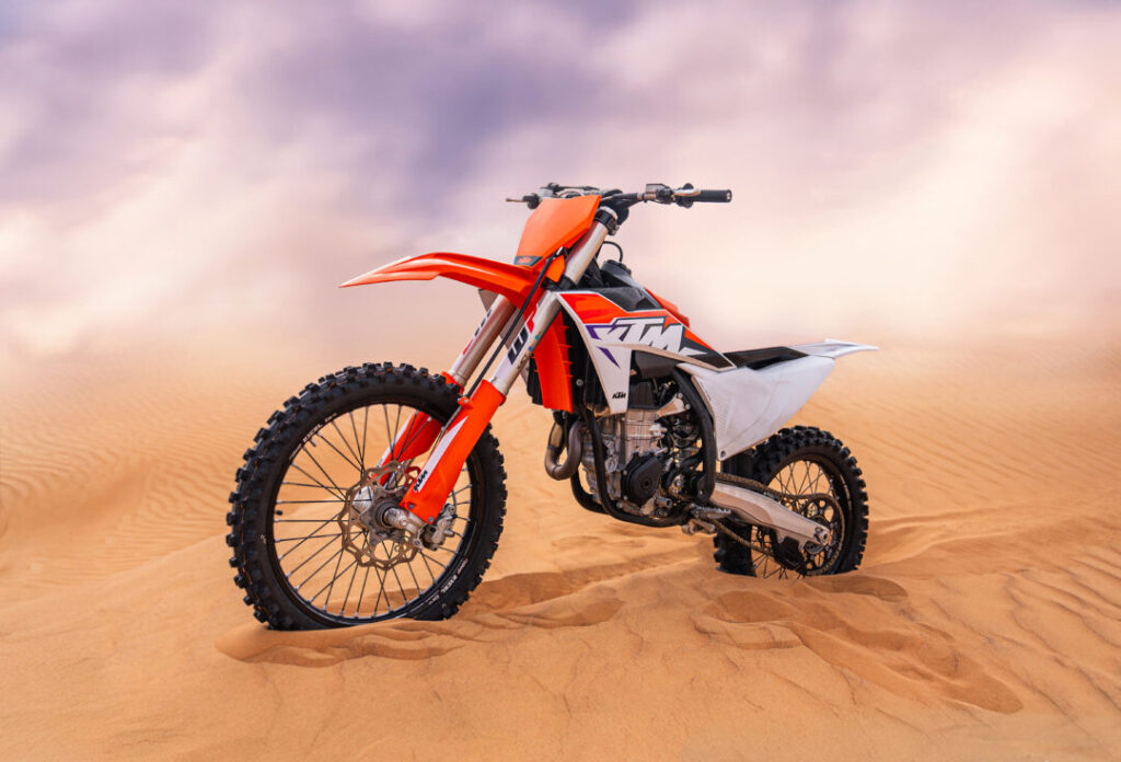Orange KTM dirt bike on the golden desert dunes of Dubai under a cloudy sky.