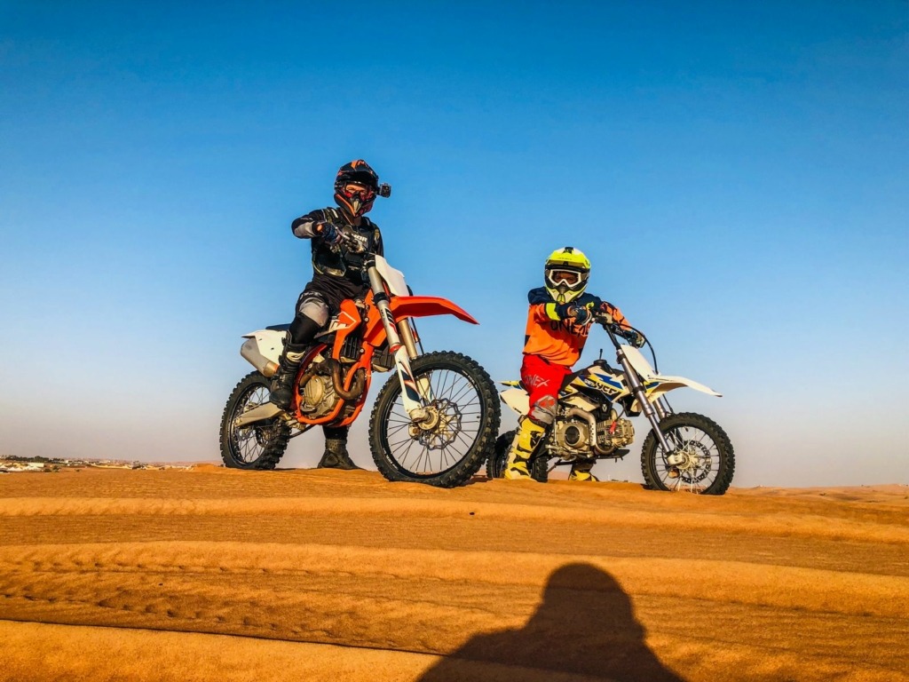 Two dirt bikers, one adult and one child, riding dirt bikes on the golden desert dunes of Dubai under a clear blue sky enjoying Dirt Bike Dubai.