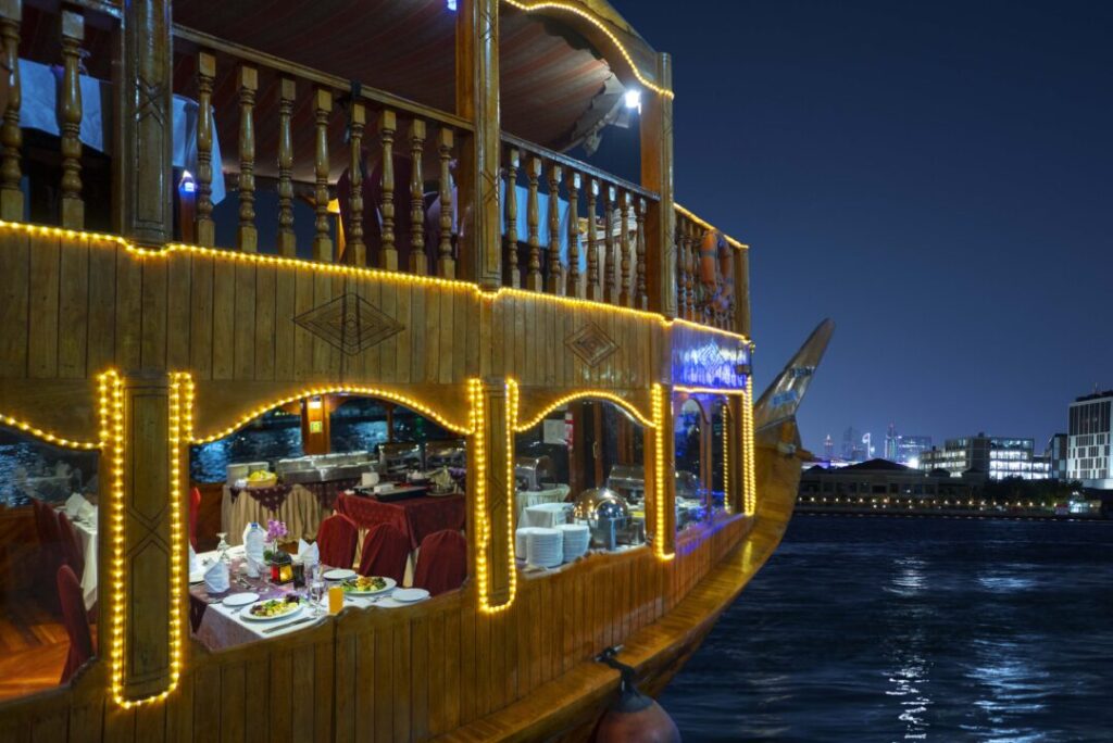 › Forever Tourism LLC A dhow cruise boat on a night-time waterfront framed artistically by an ornate archway with a hanging lantern, highlighting the Dubai Marina Dhow Cruise experience, including the popular Dubai Marina Dhow Cruise with dinner, along with details related to the Dubai Marina Dhow Cruise price and the Dubai Marina Dhow Cruise dinner price.