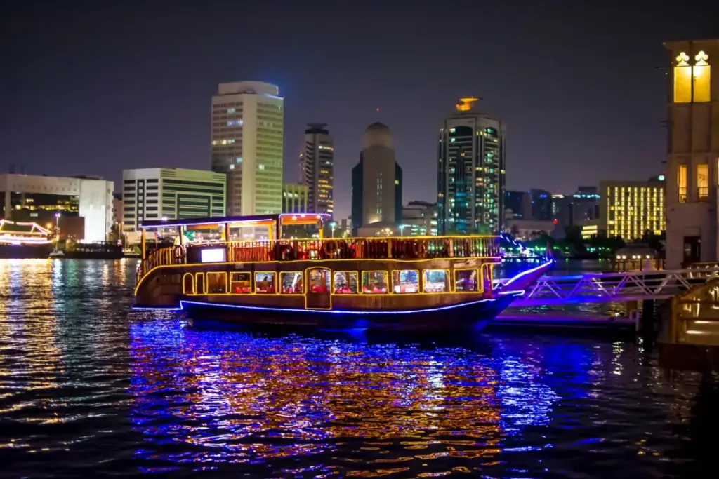 A brightly illuminated dhow cruise boat reflecting colorful lights on the water at night, with city buildings in the background, showcasing the 2 nights cruise from Dubai experience along with options like a one night cruise Dubai, a scenic Dubai to Abu Dhabi cruise, and a convenient 1 day cruise from Dubai to Abu Dhabi.