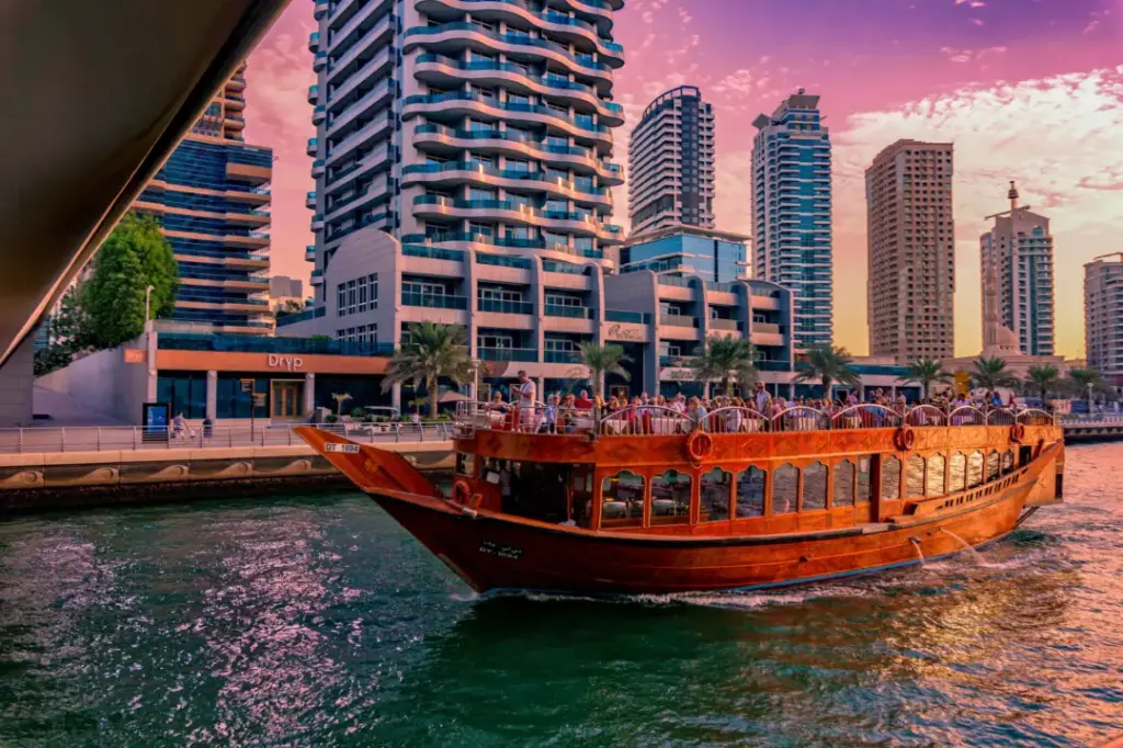 zww2bgrm1f4jiya8lt8ssd5q7qj4_1595496860_shutterstock_1292135989 › Forever Tourism LLC A wooden dhow cruise boat on calm water with a modern city skyline featuring tall towers in the background, showcasing the Dubai Marina Dhow Cruise experience, including important details such as Dubai Marina Dhow Cruise timings, the popular Dubai Marina Dhow Cruise with dinner, the overall Dubai Marina Dhow Cruise price, the Dubai Marina Dhow Cruise dinner price, and the scenic Dubai Marina Dhow Cruise location.