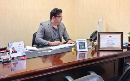 Businessman in a gray suit sits at a polished wooden desk in a quiet office, working on paperwork with a computer screen nearby.