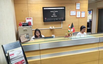 Reception desk with two women staff working on computers; wood-paneled backdrop, a wall-mounted TV displaying company info, and small flags on the counter.