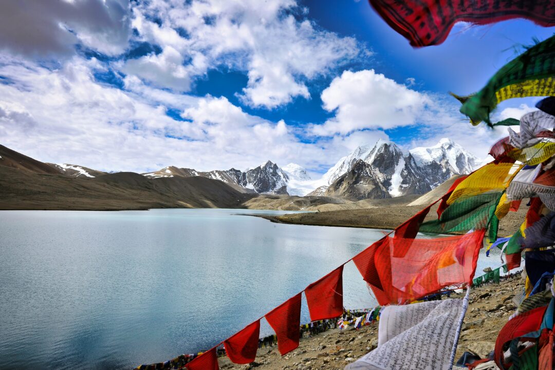 Snowy mountain range beside a turquoise lake with colorful prayer flags in the foreground.
