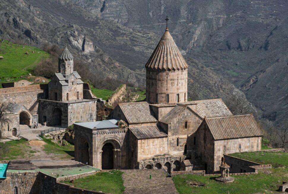 Day Trips from Yerevan: Hidden Gems, Monasteries & Scenic Escapes › Forever Tourism LLC Medieval stone church with a tall conical dome on a hillside, surrounded by rugged mountains and green slopes.