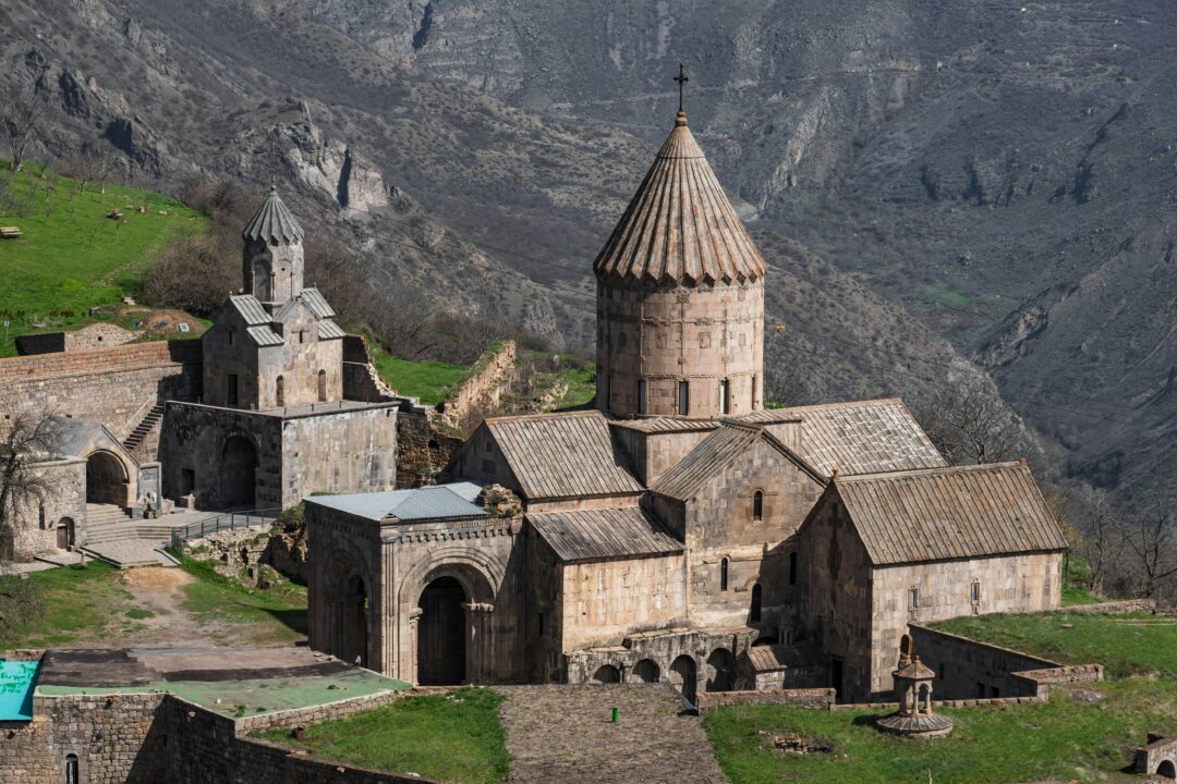 Medieval stone church with a tall conical dome on a hillside, surrounded by rugged mountains and green slopes.