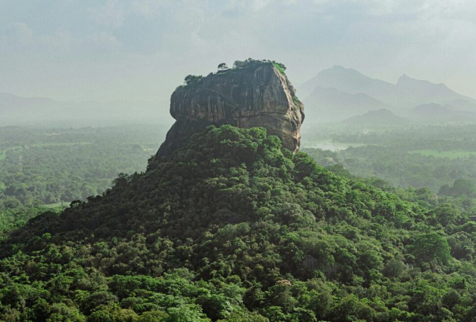 A large rocky summit rising from a green forested hill with distant mountains on the horizon.