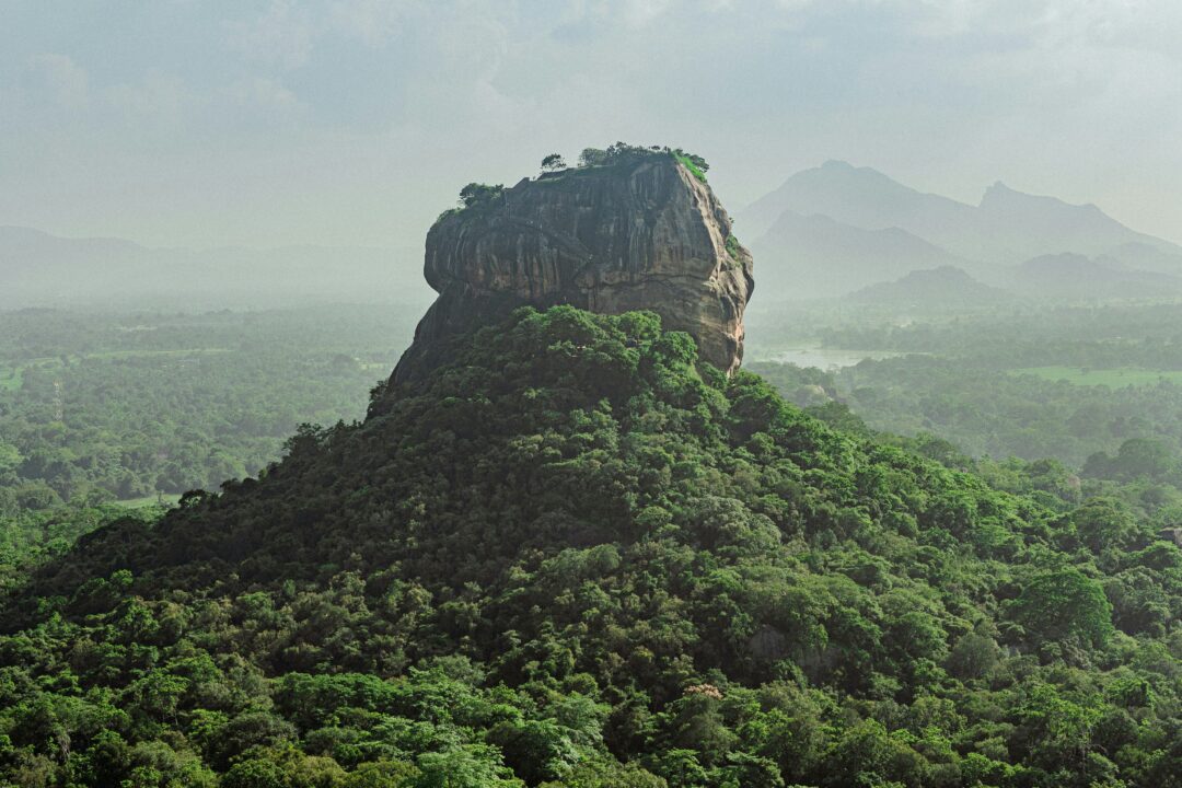 A large rocky summit rising from a green forested hill with distant mountains on the horizon.