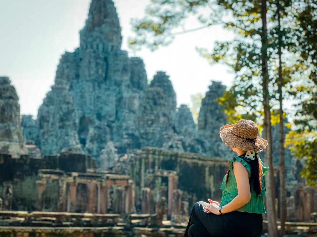 Woman in a straw hat and green sleeveless top sits on a low wall, facing ancient stone temple ruins amid trees.