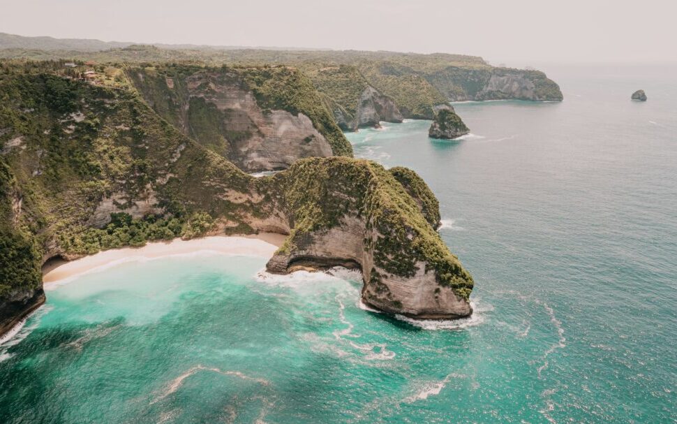 Cliffside coastline with green vegetation, a white-sand beach and turquoise sea tucked between rocky cliffs.