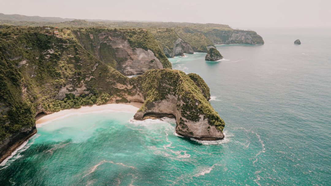 Cliffside coastline with green vegetation, a white-sand beach and turquoise sea tucked between rocky cliffs.