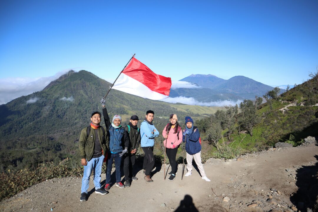 Group of hikers pose on a mountain trail; one raises a red-and-white flag with blue sky and hills in the background.