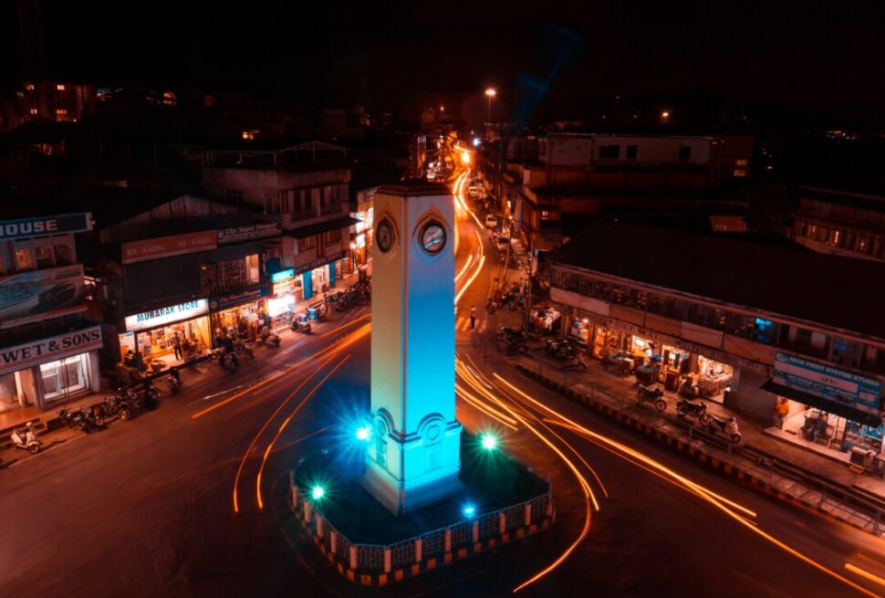 Nighttime aerial view of a roundabout with a tall, white clock tower at the center, illuminated in blue as car light trails swirl around it.