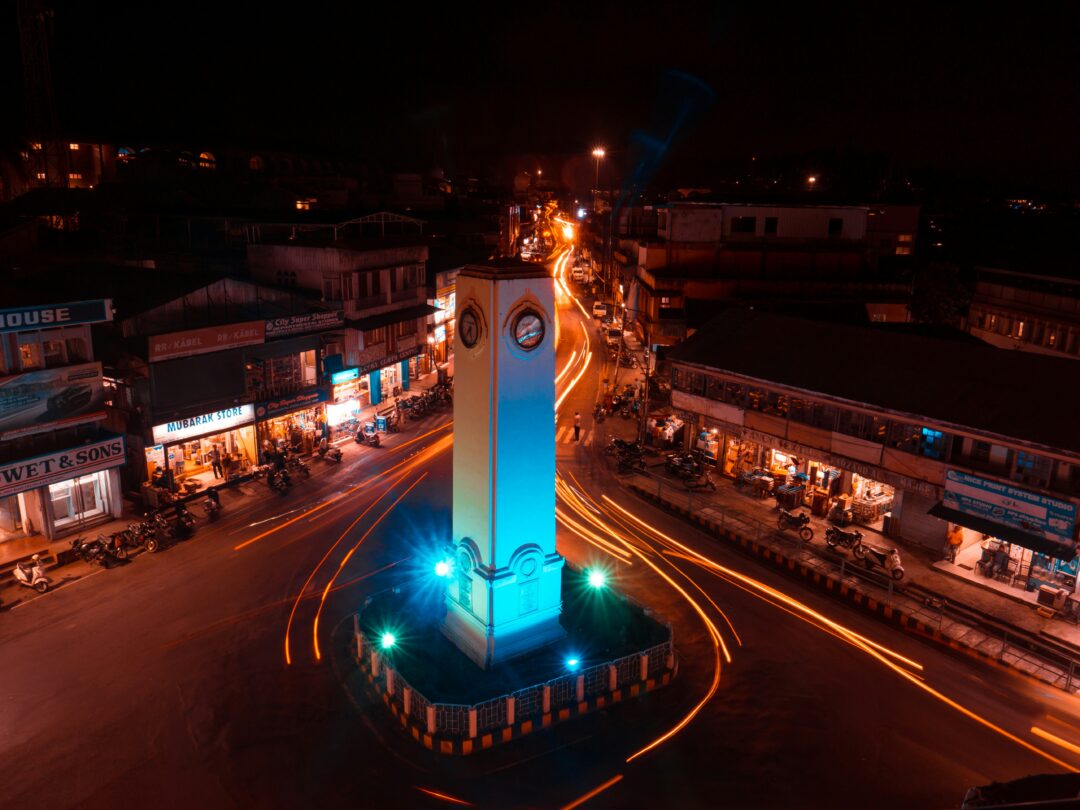 Nighttime aerial view of a roundabout with a tall, white clock tower at the center, illuminated in blue as car light trails swirl around it.