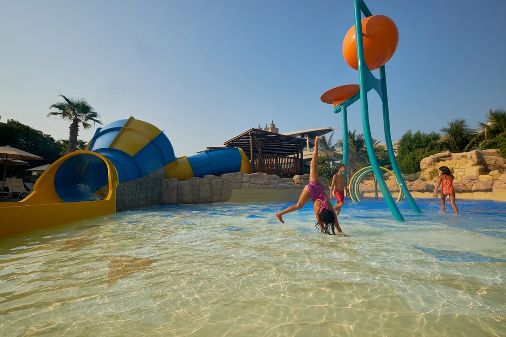 Children playing in a shallow pool at a colorful water park with blue and yellow tubular slides.