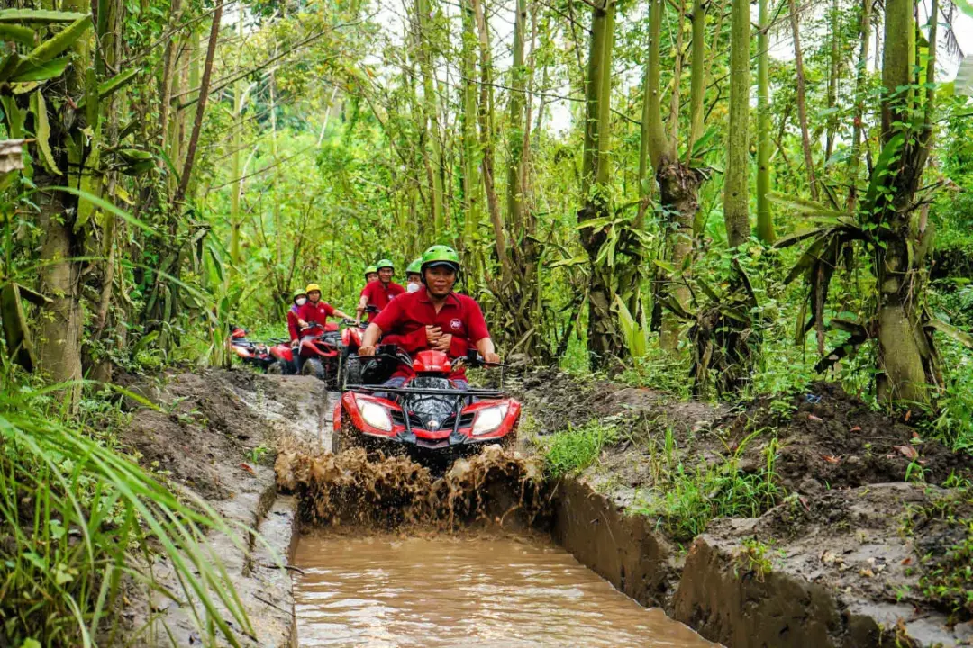 Group of people on red ATVs riding through a muddy jungle trail, splashing water as they navigate a narrow path.
