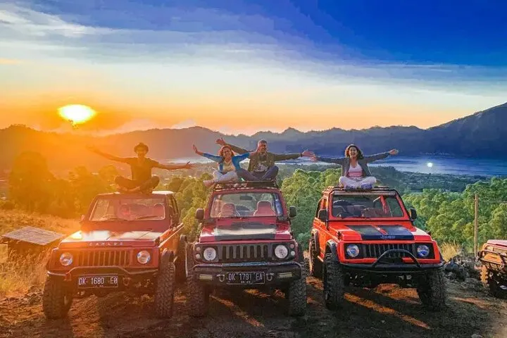 Group of people standing on three red jeeps with arms raised, sunset over mountains and valley in background, off-road adventure.