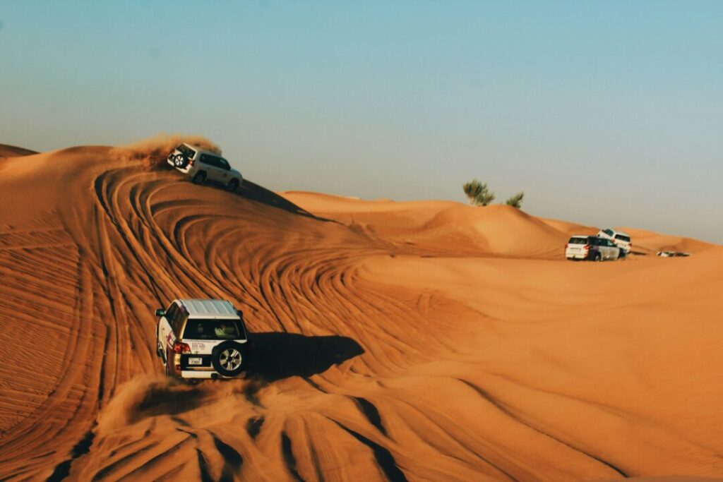SUVs climb orange desert dunes, kicking up dust under a clear blue sky (off-road convoy).