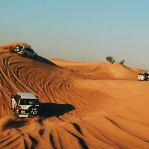 SUVs climb orange desert dunes, kicking up dust under a clear blue sky (off-road convoy).