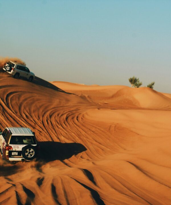 SUVs climb orange desert dunes, kicking up dust under a clear blue sky (off-road convoy).