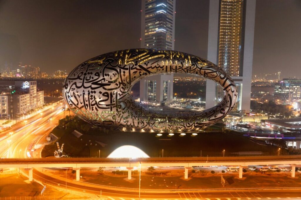 Night cityscape with a large circular Arabic calligraphy sculpture atop a hill by a freeway in Dubai's skyline.